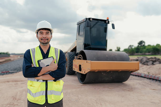 istock Confident engineer standing with arms crossed, holding tablet in front of heavy roller machine at railway construction site. 2215732828