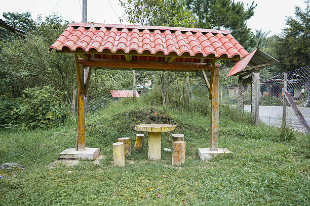 A handcrafted wooden table and stools sit beneath a red-tiled shelter, offering a peaceful and poetic corner surrounded by nature. This artistic image captures the quiet charm of rural simplicity, emphasizing natural textures, earthy tones, and a tranquil atmosphere—ideal for lifestyle editorials, design inspiration, or visual storytelling.