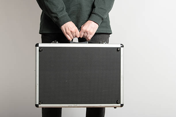 istock A person stands indoors, gripping a black briefcase with both hands. The backdrop features a plain white wall, suggesting a focus on professionalism and simplicity 2215740084