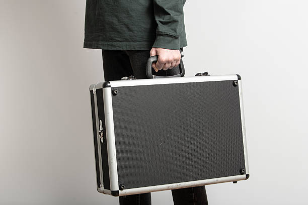 A person stands upright, holding a black case with a textured surface. The individual is dressed in casual attire, showcasing a simple and clean indoor environment with ample natural light