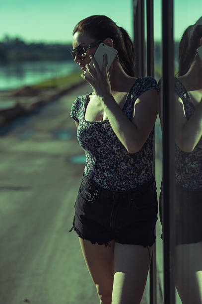 A girl in a stylish outfit is standing near a transparent glass facade, talking on the phone. A young lady wearing sunglasses looks at ease. The young woman is focused on the conversation and enjoying the warmth at the same time.