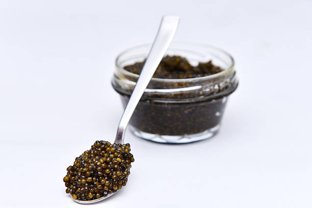 A spoon filled with delicious, fresh black caviar next to a full glass jar filled with delicacy black caviar on white background