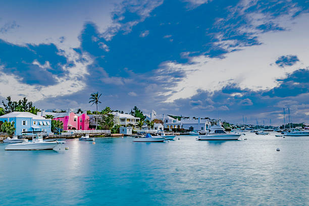 Pastel-colored houses line the waterfront under a dramatic sky with boats calmly anchored in the harbor. The serene water reflects the vivid hues of the structures and the gentle light of the setting sun at Hamilton Harbor, Bermuda.