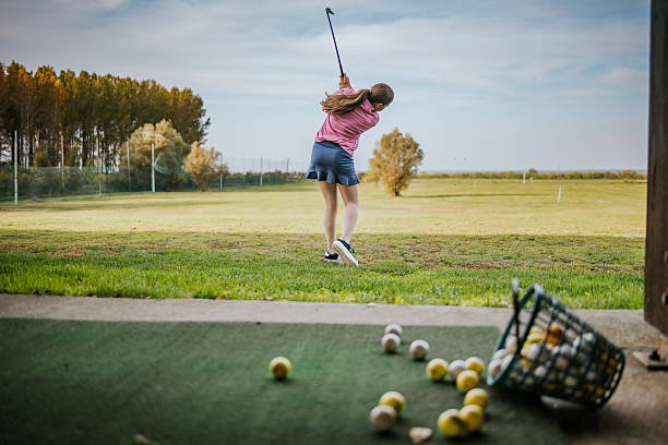 A young golfer in action at a driving range, perfecting their swing amid a scenic outdoor backdrop. Golf balls scattered in the foreground indicate active practice. This image captures the essence of golf training and the tranquility of the sport, blending focus with nature's beauty on a clear day.