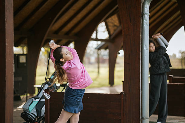 Two young golfers are practicing their swings at an outdoor driving range. The primary focus is on a girl in a pink shirt, executing a strong swing. A golf bag is visible in the background as they train in a shared space, suggesting a learning environment and dedication to improving their golfing skills.