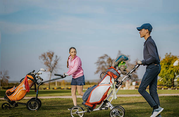 A man and a woman are seen pushing their golf bag trolleys across a lush, green course. They appear engaged and ready for a game under the clear blue sky. Their attire and focused expressions highlight the leisurely yet competitive nature of golf, capturing the essence of outdoor sportsmanship.