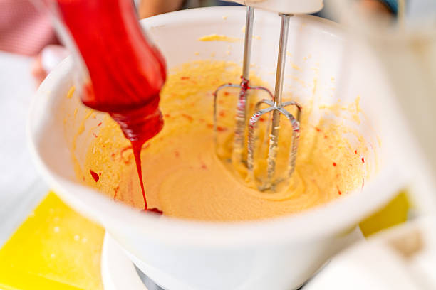 Red food coloring being added to cake batter in a white bowl with an electric mixer, creating a vibrant and colorful baking experience