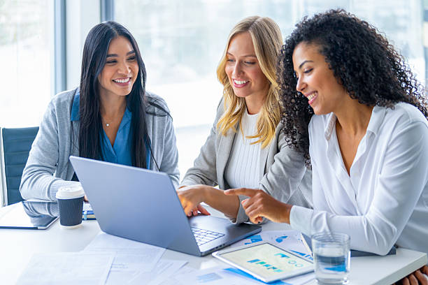 Three women meeting in a board room. There is a laptop computer on the table along with paperwork showing financial data charts and graphs