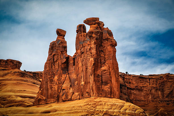 The image depicts the Three Gossips rock formation in Arches National Park, showcasing three prominent sandstone spires against a clear blue sky. The warm colors of the rock contrast with the surrounding landscape, highlighting the geological features of the area.