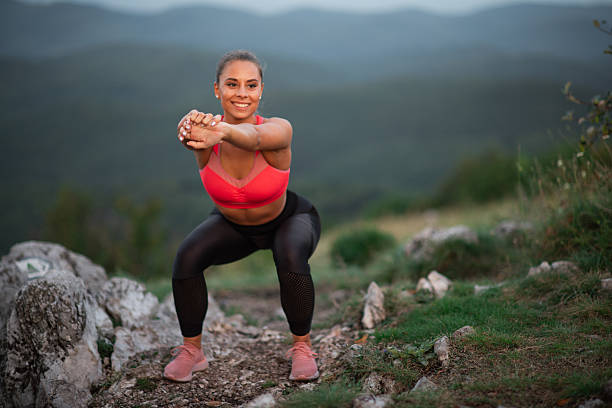 A young athletic woman in sportswear performs a squat exercise on a rocky mountain trail at sunset. She is smiling confidently, wearing a pink sports bra and black leggings, surrounded by natural scenery with rolling hills in the background. The image conveys health, vitality, and outdoor activity.