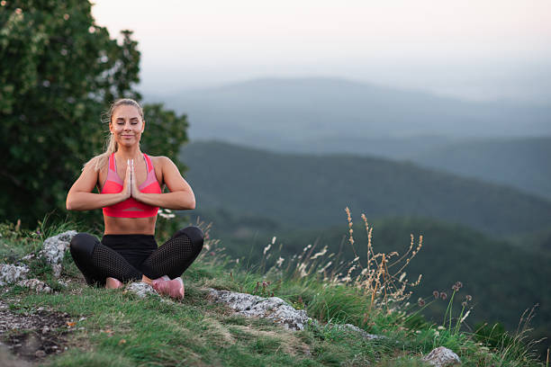 A serene young woman sits cross-legged on a grassy mountain ledge, eyes closed and hands in a prayer position. Dressed in a pink sports bra and black leggings, she meditates peacefully against a backdrop of rolling hills and a soft sky. The image conveys mindfulness, inner calm, and harmony with the natural world.