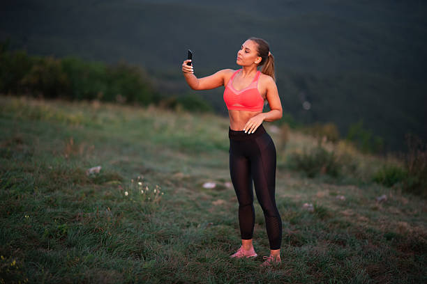 A sporty young woman in a red top and black leggings stands barefoot on a grassy hillside, holding her phone up to take a selfie. The golden-hour lighting and distant forested mountains highlight her energy and satisfaction after exercising outdoors in nature.