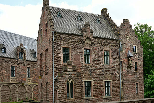 Historic Paffendorf Castle (Burg Paffendorf), located in Bergheim, North Rhine-Westphalia, Germany. This impressive structure, originally built in the 16th century, has been restored in the neo-Gothic style. Prominently featured in the foreground is a round defensive tower with crenellations, flanked by ornate windows and Gothic architectural details. The castle complex is surrounded by a moat and set within a beautifully landscaped park. Today, Paffendorf Castle serves as an event venue, museum, and visitor center for the energy company RWE