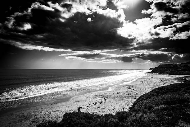 A sweeping black and white view of a beach and headland under dramatic skies. Sunlight filters through heavy clouds, illuminating the water and sand in radiant contrast. The image captures the fleeting beauty of light breaking through stormy weather.