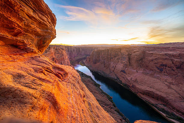 Breathtaking sunset view over the rugged cliffs of Glen Canyon with the Colorado River winding through the deep, shadowed gorge. The warm, golden light accentuates the textures and hues of the sandstone rock formations, creating a dramatic and serene desert landscape.