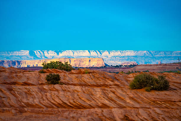 A captivating view of distant plateaus illuminated by the sun, contrasted against the rugged red rock terrain in the foreground under a vibrant blue sky. Captured near Page, Arizona, showcasing the striking geological formations of the American Southwest.