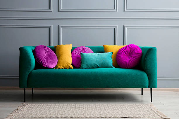 Stylish teal color sofa with different cushions near grey wall