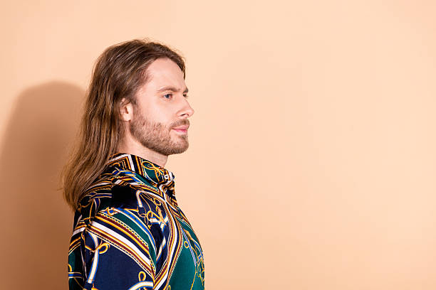 Portrait of a fashionable young man with long brown hair wearing a patterned green shirt, set against a beige background. A contemporary take on classic elegance and modern style.