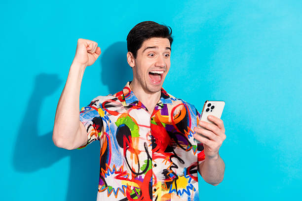istock Excited young man celebrates success while holding smartphone against vibrant blue backdrop wearing colorful shirt 2215832533