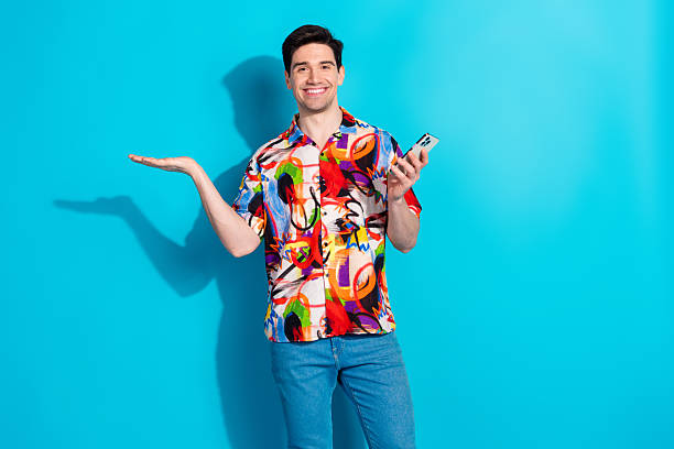 istock Young stylish man smiling brightly while holding a phone and gesturing with his hand against a blue background 2215832540