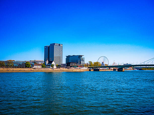 The Rhein River in Cologne, Germany, with modern architecture, a ferris wheel, and a clear blue sky—an iconic urban scene perfect for travel, skyline, and cityscape themes.