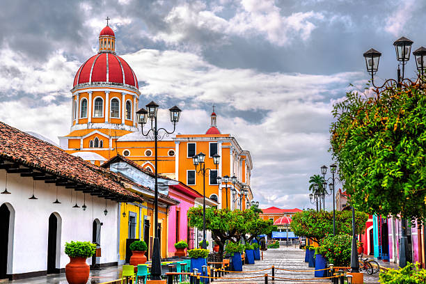 View of Granada's iconic cathedral dome rising behind a vibrant pedestrian street lined with colorful colonial buildings, trees in blue planters, and iron lampposts under a moody sky in Nicaragua.