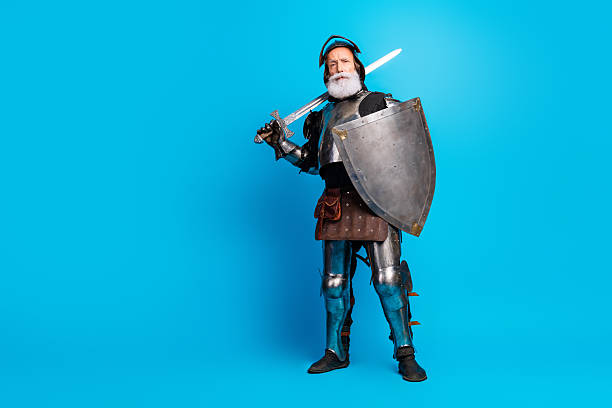 istock Elderly man dressed as a knight in medieval armor with sword and shield, standing confidently against a bright blue background 2215837656