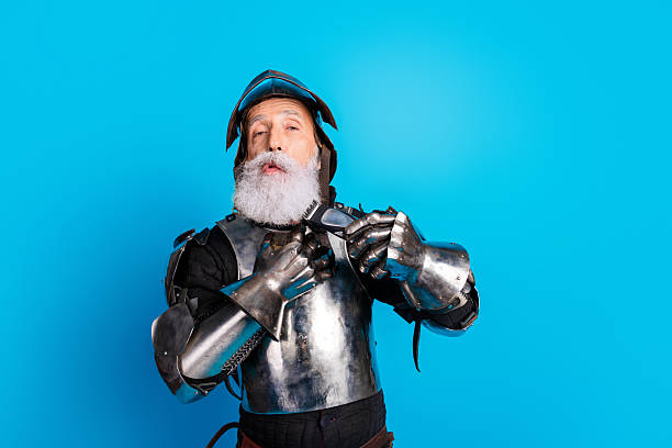istock Elderly man in medieval-style shiny armor with a steel helmet on a blue background, displaying a mix of humor and valor 2215837722