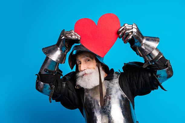 istock Charming elder dressed as a medieval knight holding a red heart on a blue backdrop 2215837723