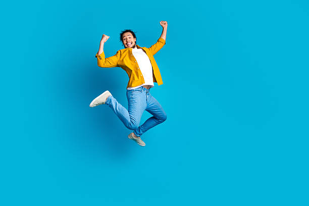 istock Young man joyfully jumping in mid air while expressing excitement on a vibrant blue background wearing casual yellow attire 2215839103
