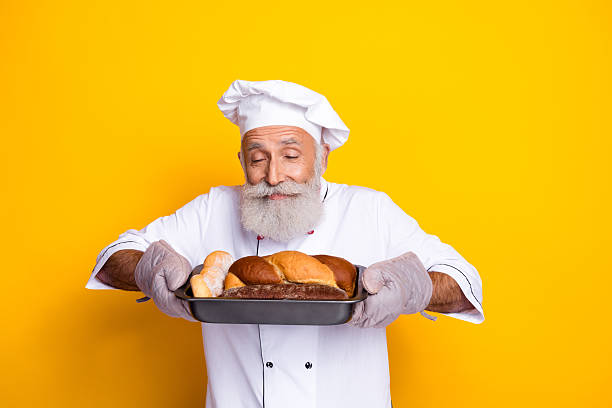 Elderly chef in a kitchen uniform showcasing freshly baked bread against a bright yellow backdrop. This image highlights culinary expertise and the joyful art of baking.