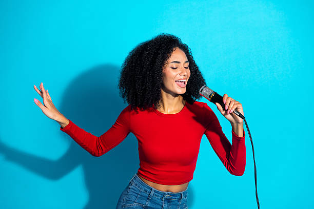 istock Young stylish woman singing passionately with a microphone on a vibrant blue background wearing a red top 2215839741