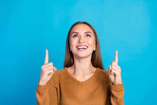istock Charming Young Woman Gesturing Upwards Against a Vibrant Blue Background, Exuding Happiness and Charm in a Stylish Brown Sweater 2215840210