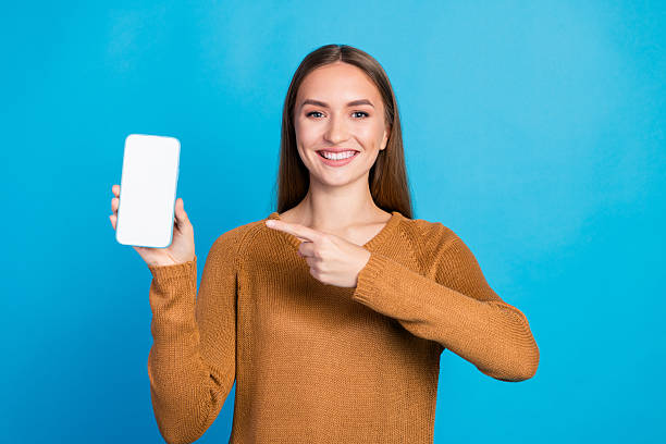 istock Smiling young woman pointing at a smartphone with a blank screen against a blue background, promoting technology and casual style 2215840230
