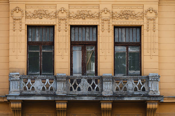 Close-up of a richly ornamented historic building featuring three tall windows with dark wooden frames and intricate stucco details. A stone balcony with elegant geometric railings adds classical charm. The warm yellow facade enhances the architectural beauty and preserved heritage.