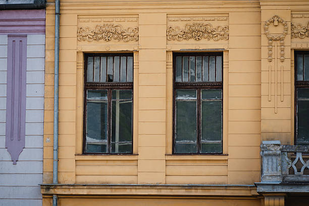 Two tall, weathered wooden windows are framed by intricate stucco decorations on a warm yellow historic building. The classical architectural style and visible aging suggest a richly layered past. The adjoining lavender-colored structure adds a touch of color contrast to the scene.