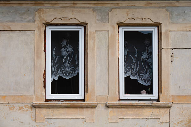 Close-up of two white-framed windows set in a faded beige facade with ornamental stucco detailing. Elegant lace curtains with floral patterns partially obscure the interior, where soft toys are visible on the windowsill. The image evokes a sense of nostalgia and quiet domestic life.