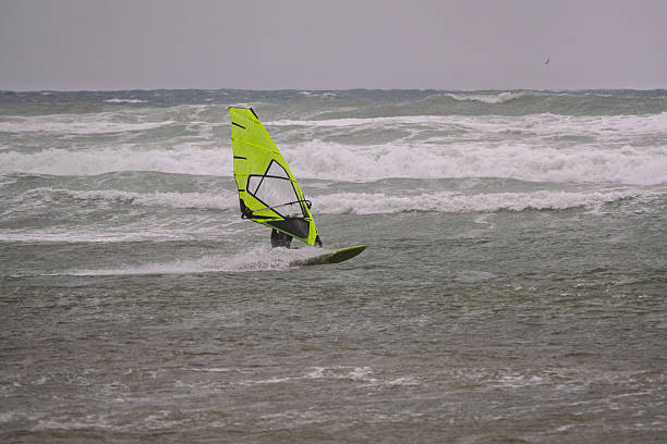 A windsurfer glides over choppy sea waters on a windy day, with another sailboarder visible in the distance. Captured along the Tuscan coastline, Italy.