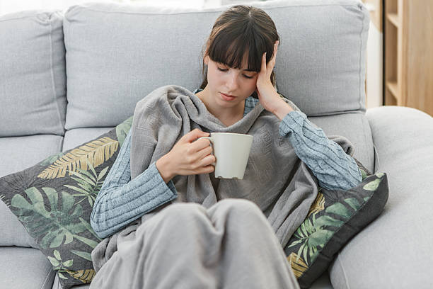 istock Sick woman sitting on a couch, wrapped in a blanket, holding a mug 2215848349