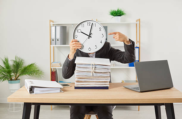 istock Funny worker employee hiding head behind the clock at workplace in office with a pile of folders. 2215856285