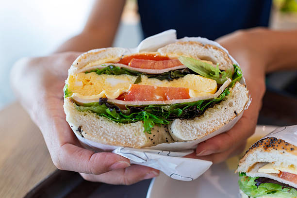 Close-up of a freshly made bagel sandwich filled with egg, cheese, lettuce, and tomato, held in both hands, showcasing a healthy and delicious meal option.