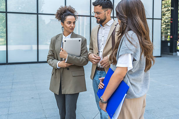 istock Diverse team of corporate professionals in formal attire collaborating outdoors, engaged in strategic discussion while analyzing data on laptop in front of modern glass office building. 2215870266