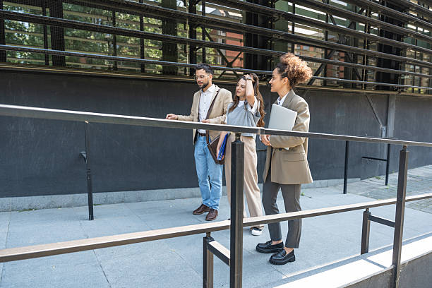 istock Diverse team of corporate professionals in formal attire collaborating outdoors, engaged in strategic discussion while analyzing data on laptop in front of modern glass office building. 2215870339