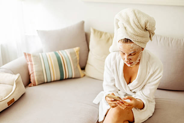 istock Woman Relaxing With Facial Mask While Using Phone on Comfortable Couch 2215878629