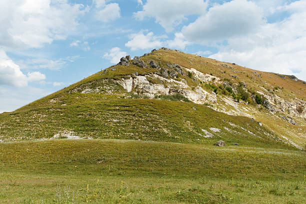 Rolling green hill with rocky outcrops and sparse vegetation, natural landscape style, under a cloudy blue sky, concept of peaceful wilderness