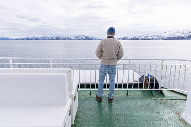 Man on ferry deck admiring expansive snowy mountains. Scenic winter travel across icy waters. Solitary figure enjoying quiet, natural beauty in cold climate.