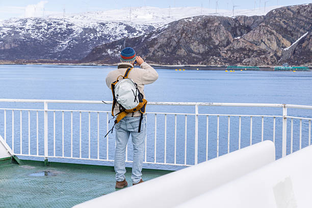 Person in warm clothing on ferry deck gazing at mountains. Scenic view of snowy peaks and calm blue waters. Winter travel and exploration theme outdoors.