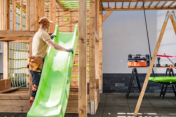 A skilled construction worker is seen positioning a vibrant green slide onto a sturdy wooden playset. The activity takes place in a well-lit residential backyard, surrounded by lightly shaded structures. Tools and equipment are organized nearby, highlighting a focused work environment.