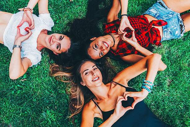 Three young women relax on green grass, forming heart shapes with their hands and enjoying a carefree summer day together. Their colorful outfits and playful poses capture the spirit of friendship, joy, and connection in an outdoor setting.