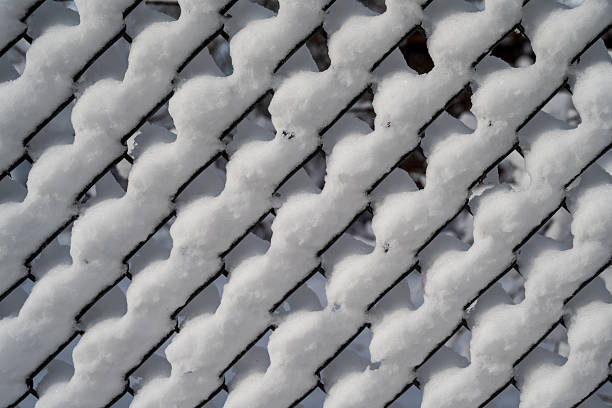 A small metal structure with a triangular roof sits covered in a thick layer of snow, enclosing an open pit. Thin vertical bars give it a cage-like appearance, contrasting with the surrounding snowy landscape. Behind it, a brick building with barred windows adds an urban touch, while trees and snow-covered ground create a winter atmosphere. The scene captures a quiet, cold day in a residential or industrial area.